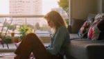 Mulher em um apartamento sentada no chão encostada no sofá de cabeça baixa